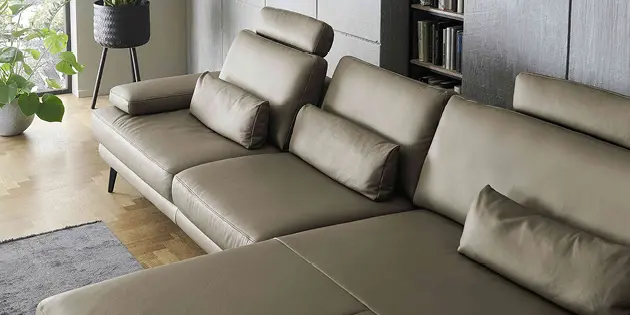 Detailaufnahme eines modernen olivgrünen Ledersofas mit verstellbaren Kopfstützen und passenden Rückenkissen. Schwarze Metallfüße, Parkettboden, graue Wand und Pflanze am Fenster unterstreichen das elegante Wohnambiente.